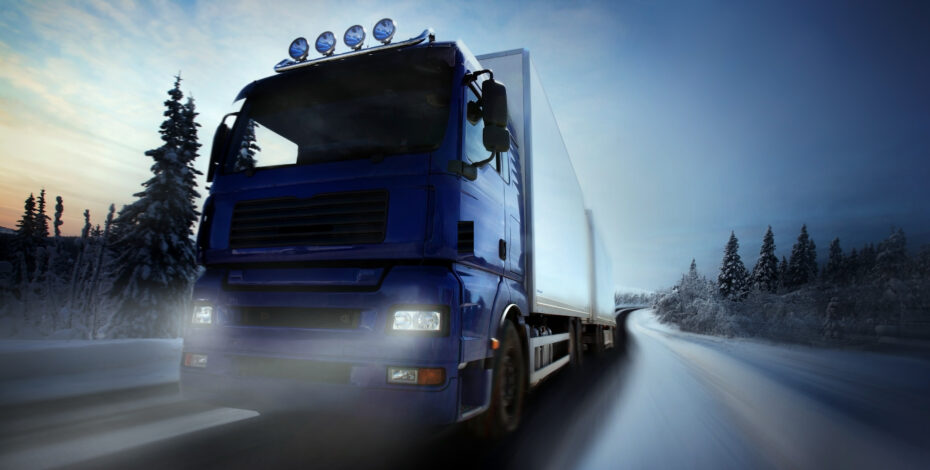 A blue semi-truck driving on a snowy road with headlights on, surrounded by snow-covered trees under a dusky sky.